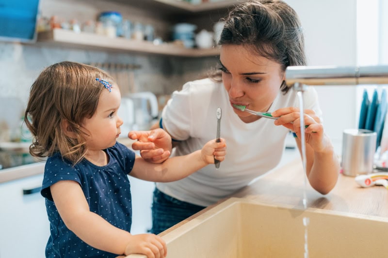 Mom teaching tolder to brush teeth at sink using filtered water system.